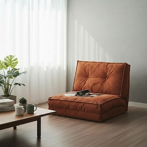 Puede incluir: Una silla de suelo de pana naranja con respaldo y asiento acolchados. Una revista descansa sobre el asiento. La silla está en una habitación con suelo de madera, una cortina blanca y una pequeña mesa de madera.