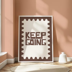 May include: A framed print with the words "KEEP GOING" in brown block letters. The print has a white background and a brown border with a scalloped edge. The frame is white, and the print is leaning against a peach-colored wall.