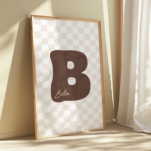 May include: A framed art print featuring a large, brown letter "B" with two white circles and the name "Billie" in a cursive font. The background has a checkered pattern in beige and white. The frame is a light brown color.