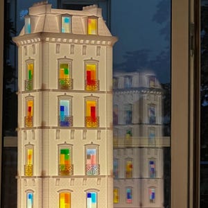 Könnte beinhalten: Eine weiße, mehrstöckige Gebäude-Lampe mit bunten Buntglasfenstern. Die Lampe hat eine detaillierte Fassade mit Balkonen und einem Dach. Die Fenster sind mit verschiedenen Farben beleuchtet und erzeugen ein warmes Leuchten.