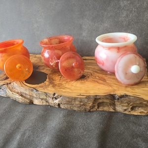 Op de afbeelding: Drie kleine, ronde potjes met deksel van hars of glas. Eén potje is feloranje, één is gemarmerd rood-oranje en de derde is gemarmerd roze en wit. Elk potje heeft een bijpassend afneembaar deksel met een kleine knop als handvat.