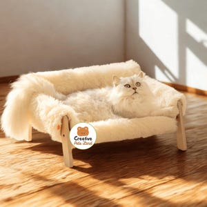 May include: A fluffy, cream-colored cat rests in a plush, cream-colored pet bed. The bed has a wooden frame and is covered in soft, faux fur. The bed is labeled with the text "Creative Pets Land".