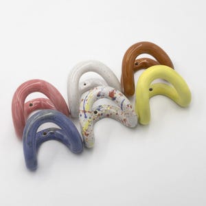 Op de afbeelding: Een verzameling van zes boogvormige wandhaken van keramiek in verschillende kleuren en afwerkingen. De haken zijn voorzien van effen roze, blauwe, gele en bruine glazuren, evenals een witte gespikkelde afwerking en een witte basis met kleurrijke verfspatten.