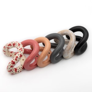 Könnte beinhalten: Ein Set aus sechs geschwungenen, skulpturalen Ringen, die in einer Reihe angeordnet sind. Die Ringe haben verschiedene Oberflächen, darunter eine cremefarbene Basis mit roten und schwarzen Sprenkeln, mattes Rot, Beige, Dunkelgrau, Marmorbeige und Schwarz.