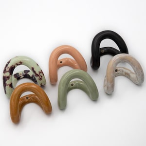 Op de afbeelding: Een verzameling van zes boogvormige keramische wandhaken in diverse kleuren en afwerkingen, waaronder effen zwart, glanzend perzik, saliegroen, mosterdgeel, gespikkeld lichtgroen en gemarmerd beige, elk voorzien van een enkel montagegat.