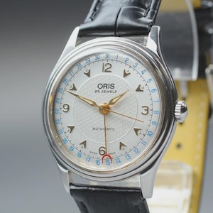 May include: An Oris automatic wristwatch with a silver textured dial, gold-tone hands, and a date pointer hand with a red crescent tip. The watch features a silver case, a black leather strap, and a blue outer ring displaying the numbers 1 through 31.