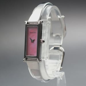 Puede incluir: Un reloj Gucci plateado con una esfera rectangular rosa. El reloj tiene una correa plateada y agujas negras. La palabra "GUCCI" está impresa en la esfera. El reloj se muestra sobre un soporte transparente.