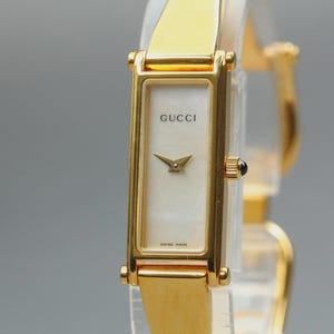 Puede incluir: Un reloj de pulsera Gucci dorado con una esfera rectangular y una esfera blanca. El reloj tiene una correa dorada y una pequeña corona negra. La esfera muestra el logotipo de Gucci y las palabras "Swiss Made".