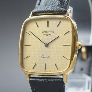 Könnte beinhalten: Eine goldfarbene Longines Quartz-Uhr mit quadratischem Zifferblatt und schwarzen Zeigern und Markierungen. Die Uhr hat ein schwarzes Lederarmband. Das Zifferblatt zeigt das Longines-Logo und das Wort "Quartz".