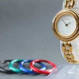 May include: A gold-tone Gucci wristwatch with a round white face, thin black hands, and a link bracelet. The watch is displayed on a clear stand, with five interchangeable circular bezels in silver, black, green, blue, and red arranged below.