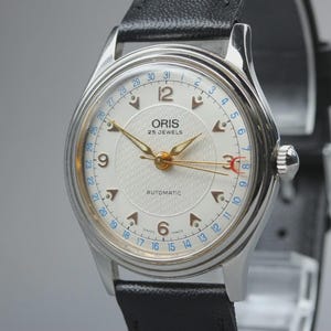 Puede incluir: Un reloj de pulsera automático Oris con caja plateada y correa de cuero negro. La esfera blanca presenta manecillas doradas, números arábigos en el 6 y el 12, marcadores de hora triangulares y un anillo de fecha azul con un puntero rojo.