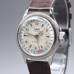 May include: An Oris automatic wristwatch with a silver-toned circular case, a textured cream-colored dial, and Arabic numerals. It features a date pointer hand with a red crescent tip, a fluted bezel, and a dark brown textured leather strap.
