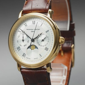 May include: A gold-tone Frederique Constant Geneve wristwatch with a white textured dial, Roman numeral hour markers, and a brown leather strap. The watch features a moon phase indicator, day and date sub-dials, and black hands.