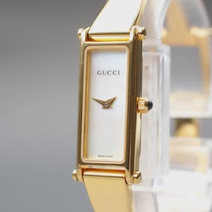 Puede incluir: Un reloj de pulsera rectangular en tono dorado con esfera blanca, el logotipo de Gucci y manecillas doradas. El reloj tiene una correa de metal pulido en tono dorado y una pequeña corona en el lateral. El texto Gucci y Swiss Made es visible.