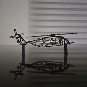 May include: A black metal helicopter model with intricate cutouts, displayed on a dark surface. The model features a main rotor, tail rotor, and fuselage, with a clear reflection beneath. The background is a neutral, light-toned wall.