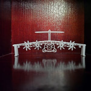 Peut inclure: Objet décoratif blanc en forme d'hélicoptère, avec un corps central et quatre motifs en forme d'hélice de chaque côté. L'objet a une base plate avec deux poteaux de support. Le fond est rouge foncé.