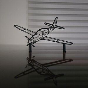 May include: A black metal airplane sculpture with a propeller and wings, set against a neutral background. The sculpture is reflected on a glossy surface, creating a mirrored effect. The design is a detailed outline of an aircraft.