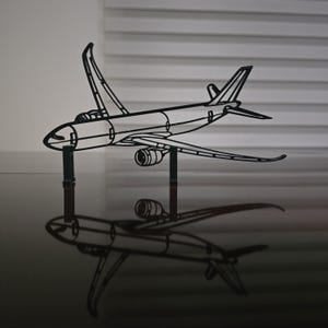 May include: A black metal airplane model with detailed wings, tail, and engine components. The model is mounted on two black supports, casting a reflection on the glossy surface below. The design is a minimalist representation of an aircraft.
