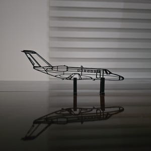 May include: A black wireframe model of a jet airplane, standing on two black supports. The model's design includes wings, a tail, and windows. The jet's reflection is visible on the glossy black surface below.