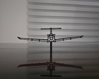 May include: A black metal airplane model with a propeller and wings. The airplane is standing upright on a black surface, and its reflection is visible. The background is a white wall and a window with horizontal blinds.