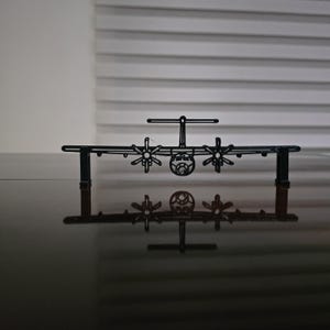 May include: A black metal airplane model with four propellers, displayed on a reflective surface. The model is a detailed silhouette of a cargo plane, showcasing the wings, fuselage, and tail. The image is a front view of the model.