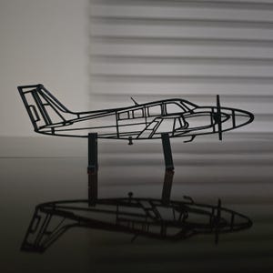 May include: A black metal airplane sculpture with a propeller and a detailed frame. The sculpture is supported by two dark green stands, casting a reflection on the glossy surface below. The design captures the essence of an aircraft.