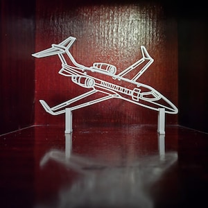 May include: A white, stylized airplane model with detailed outlines of the fuselage, wings, and tail. The model is supported by two small, rectangular stands. The background is a dark red-brown color.