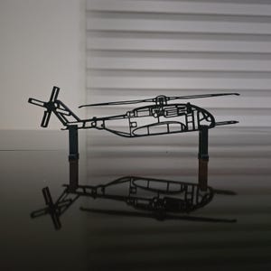 May include: A black metal helicopter model with detailed cutouts, including the rotor blades and tail. The model is mounted on two vertical supports, casting a reflection on the glossy surface below. The design showcases the helicopter's structure.