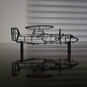 May include: A black metal sculpture of an airplane, featuring a distinctive radar dome and propeller details. The sculpture is mounted on two vertical supports, casting a reflection on the glossy surface below. The design is a detailed wireframe.
