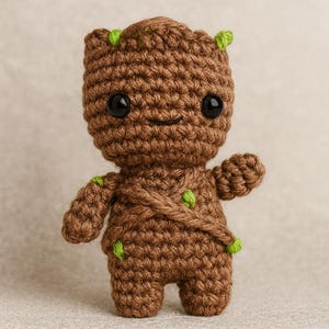Puede incluir: Una figura de ganchillo marrón hecha a mano de un personaje con grandes ojos negros y una pequeña sonrisa. La figura tiene detalles verdes en la cabeza y el cuerpo, y posa con un brazo levantado. La figura mide aproximadamente 15 cm de alto.