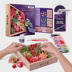 Puede incluir: Un kit de manualidades para crear suculentas de arcilla. El kit incluye un marco de madera, pinturas, herramientas e instrucciones. La caja dice "Wood & Craft Succulents" y "Sensory Art". También se ve un cartel de "No molestar".