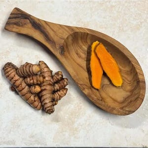 Peut inclure: Une cuillère de service en bois contient deux tranches de curcuma orange vif. Un tas de racines de curcuma fraîches se trouve à côté de la cuillère. La cuillère et le curcuma sont sur une surface de couleur claire. La cuillère est en bois avec un grain naturel.