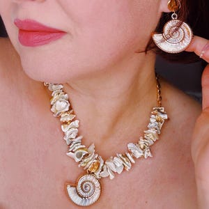 Peut inclure: Un ensemble de bijoux en coquillages comprenant un collier et des boucles d'oreilles. Le collier présente un grand pendentif en forme de spirale et est orné de divers fragments de coquillages. Les boucles d'oreilles reprennent le design du pendentif. Les bijoux ont des accents dorés.