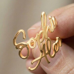 Può includere: Un anello dorato con il nome "Sophia Ala" scritto in corsivo. L'anello è tenuto tra le dita, evidenziando il design personalizzato. La scrittura elegante dell'anello è visibile, mostrando il design fluido.