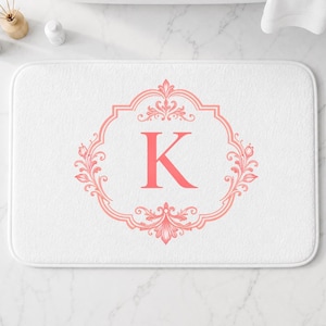 May include: A rectangular white bath mat featuring a centered coral-colored letter K enclosed within an ornate, symmetrical floral frame. The design is printed in a soft coral hue, providing a clear contrast against the plain white fabric surface.