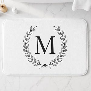 May include: A rectangular white bath mat featuring a centered black letter M enclosed within a symmetrical black line-art laurel wreath design. The mat has rounded corners and a soft, textured surface, providing a clean and simple aesthetic.