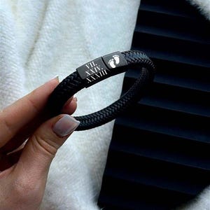 Puede incluir: Pulsera de cuero trenzado negro con cierre rectangular. El cierre presenta un pequeño símbolo de huella y números romanos. La pulsera se sostiene contra un fondo blanco y difuso y una superficie negra texturizada.
