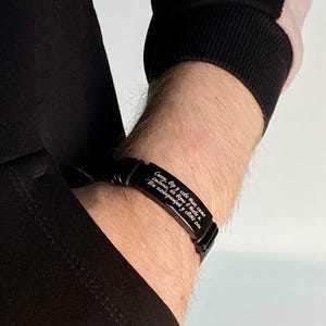 Puede incluir: Pulsera negra con texto grabado en blanco. La pulsera se lleva en la muñeca, con una camiseta negra de manga larga visible. El texto de la pulsera está en un idioma extranjero.