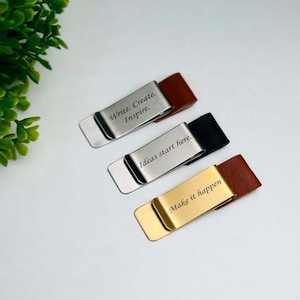 Puede incluir: Tres clips de dinero de metal con detalles de cuero. Uno es dorado con "Make it happen" grabado, otro es plateado con "Ideas start here", y el último es plateado con "Write. Create. Inspire."