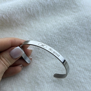 May include: A silver-colored cuff bracelet with the words "do good" and an infinity symbol engraved on the outside. The bracelet is curved and open-ended. The background is a soft, white, textured surface.