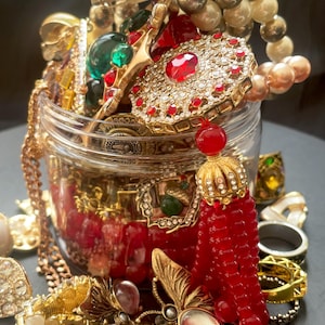 Pode incluir: Um frasco de vidro transparente transbordando com uma variedade de joias vintage. As joias incluem colares, pulseiras, anéis e broches dourados, adornados com pérolas, pedras preciosas vermelhas e verdes e strass. Uma borla vermelha de contas pende do frasco.
