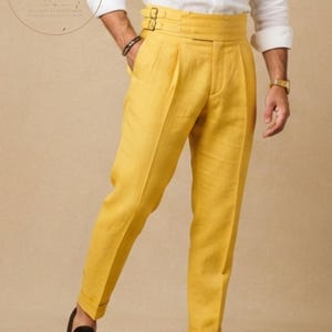 May include: Bright yellow high-waisted trousers featuring a wide waistband with two side buckle adjusters, front pleats, and cuffed hems. The trousers are paired with a white long-sleeved shirt and dark brown loafers, with a ZstyAtelier logo visible.