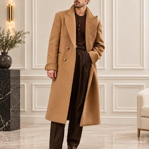 May include: A long camel-colored wool overcoat with wide lapels and double-breasted button closure, worn over a dark brown henley shirt and dark brown pleated trousers with a matching leather belt and brown leather dress shoes.