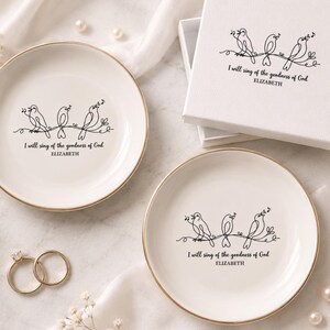 Può includere: Due piattini rotondi in ceramica bianca con bordi dorati, ciascuno con un'illustrazione a linee nere di tre uccelli su un ramo e il testo "I will sing of the goodness of God" sopra il nome "ELIZABETH" in una scatola regalo bianca.