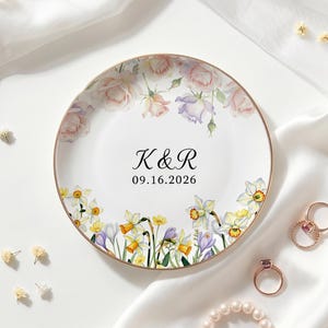 Puede incluir: Un plato redondo de cerámica blanca con borde dorado, que presenta un diseño floral en acuarela de rosas rosadas en la parte superior y narcisos amarillos y azafranes morados en la inferior. El centro muestra el texto negro K&R y la fecha 16.09.2026.