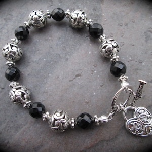 May include: A silver bracelet with black beads and a heart-shaped charm. The charm has a keyhole design and is attached to a toggle clasp.