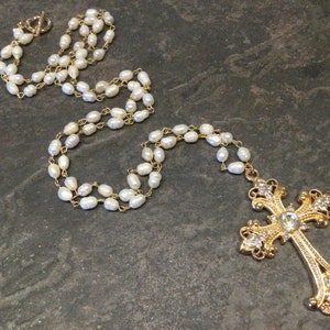 May include: A long pearl necklace with a gold ornate cross pendant featuring clear rhinestones. The necklace is on a gray textured surface.