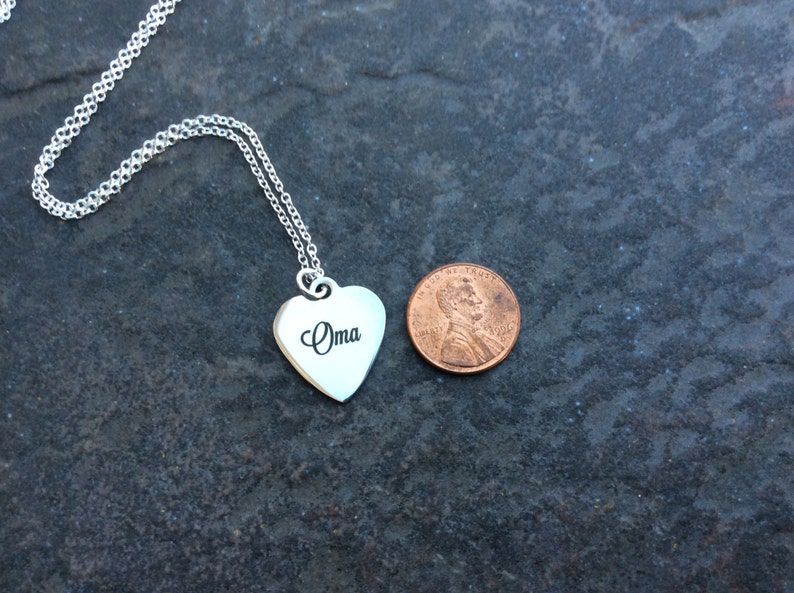 May include: A silver heart-shaped pendant necklace with the word " Oma" engraved on it. The pendant is about the size of a US penny.