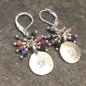May include: Silver earrings with a round charm featuring a floral design. The charms are adorned with a cluster of colorful gemstones, including pink, red, blue, and gray.