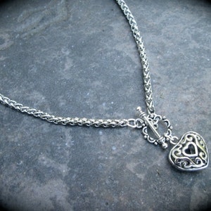 May include: A silver chain necklace with a heart-shaped pendant. The pendant has a detailed design and a small keyhole in the center.